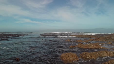 Drone-flying-low-and-fast-between-boulders-onto-sea-front-with-bamboo-covered-waters-as-waves-come-in-the-perspective-rises-displaying-open-ocean-on-a-partly-cloudy-day