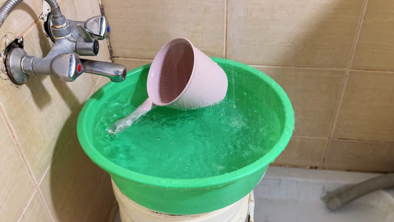 Close up of a running bathroom faucet filling a green plastic basin with water while a cup overflows inside, highlighting household water usage and indoor plumbing environment