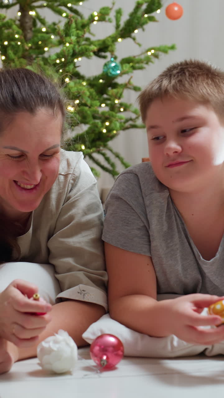 Madre e figlio bianchi che giocano con decorazioni sotto l'albero di Natale, mani che sistemano palline sul pavimento con risatine e attenzione curiosa, scena di decorazione del soggiorno che mostra la madre come insegnante
