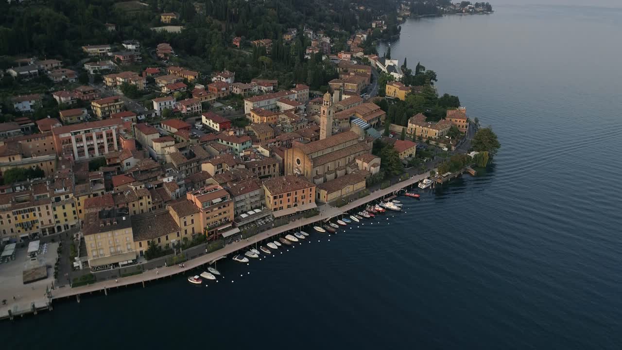 Ripresa aerea con drone della città sul Lago di Como, in Italia, che mostra edifici storici, il campanile della chiesa, il porticciolo con le barche e le acque calme del lago, creando una vista panoramica da cartolina europea.