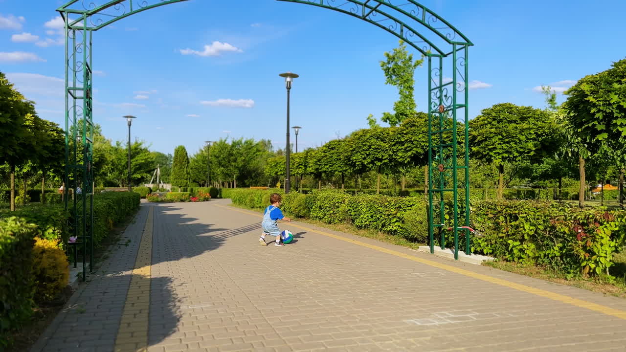 Cute little boy in jeans romper picks up a ball and runs with it. Lovely kid having fun in the sunny green park.