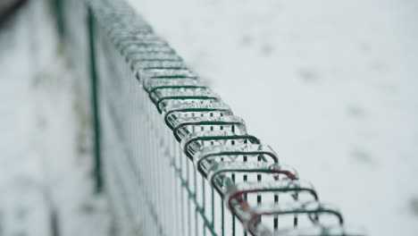 close-up of green metal fence coated with ice, showcasing icicles hanging from the wires against a blurred snowy park background, emphasizing frozen textures and the stark coldness of winter