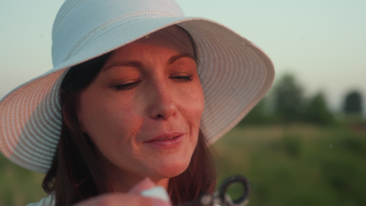 Close up of tourist wearing white sun hat gently blowing bubble outdoors during golden hour, framed by soft evening light and lush green backdrop