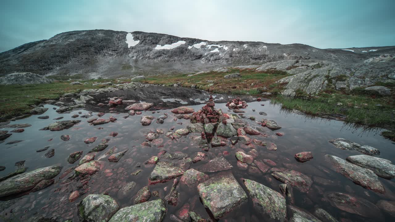 A cairn, assembled from colorful rocks, stands amidst a shallow stream in a mountainous region