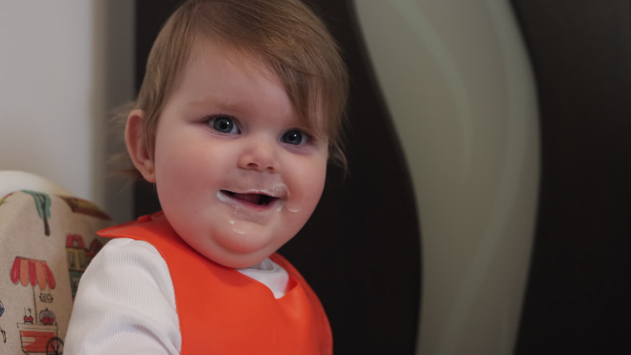 Cheerful baby girl with messy face takes break from eating, looks at camera