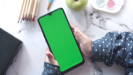 child holding smartphone with green screen, surrounded by school supplies.