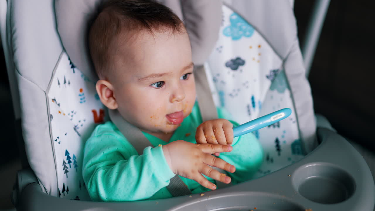 Sweet little kid plays with a food after being fed. Cute baby sitting in a chair takes a spoon into mouth. Close up.