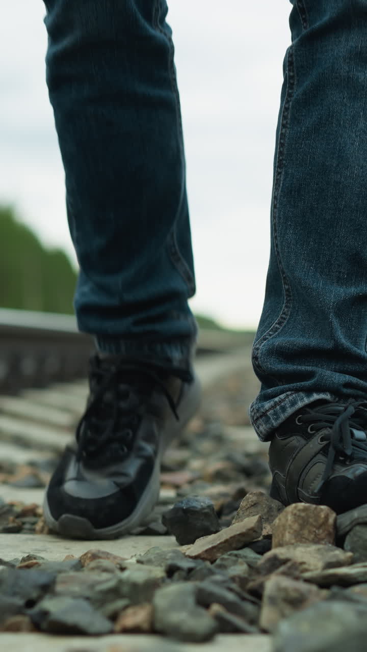 vista cercana de la pierna de alguien con pantalones vaqueros y zapatos de lona, caminando cansado en un ferrocarril con piedras, el fondo incluye un borrón de árboles y postes eléctricos