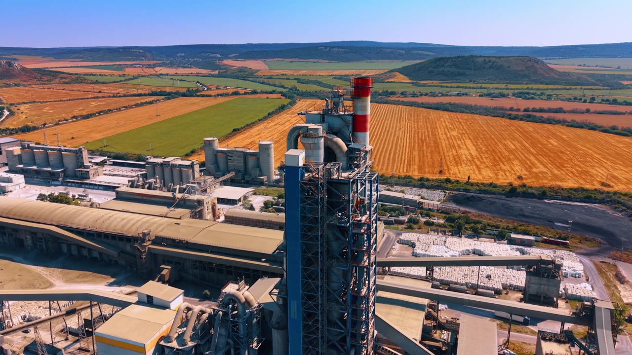Cement plant tower and silos surrounded by farmland. Oblique drone view shows a tall processing tower, red chimney, conveyors, silos and harvested fields beyond the site.
