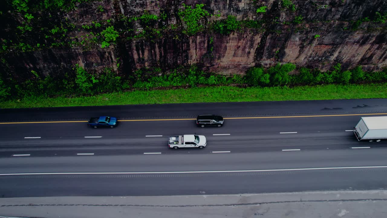 Top-down aerial of a stepdeck semi truck hauling construction equipment along a cliffside highway.