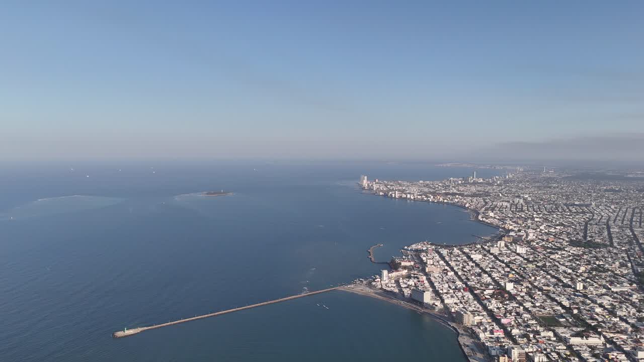 Wide aerial view of a coastal city, featuring still waters, a long breakwater, and a distant island under a clear sky.