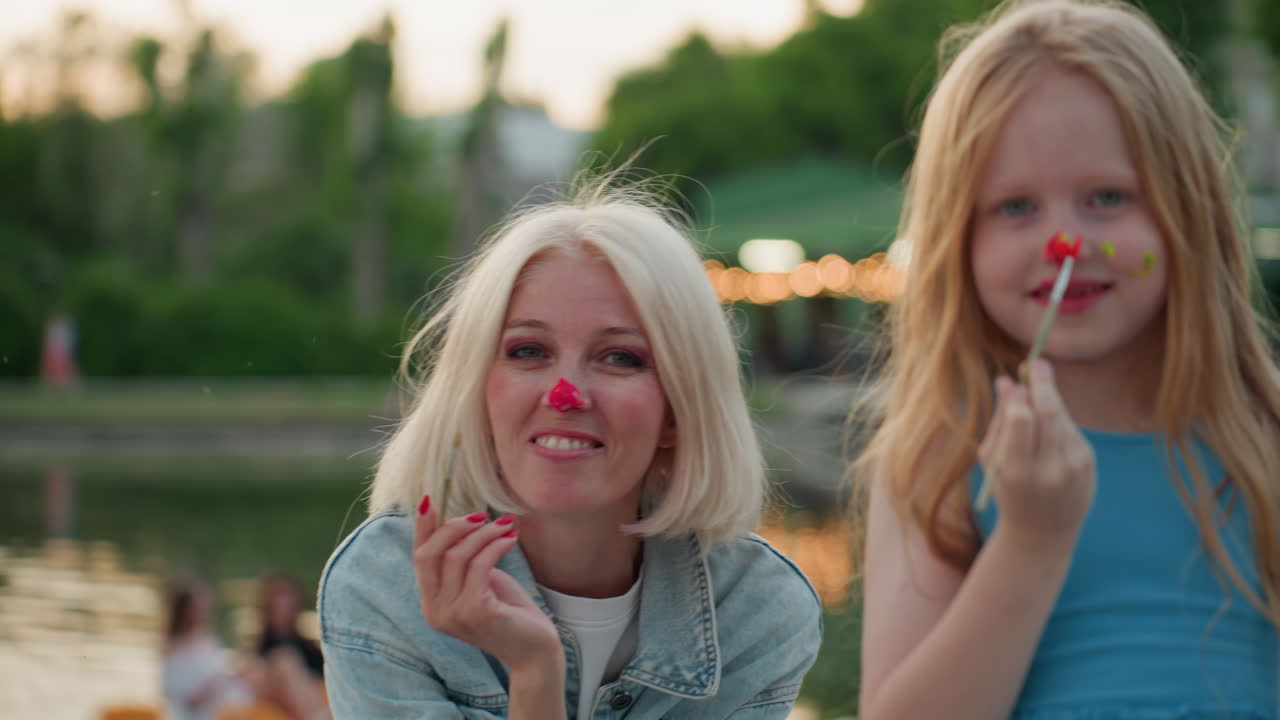 portrait of smiling mother and daughter by lake during sunset painting faces with bright paint using brushes, joyful moment with colorful splatters on skin and denim jacket in soft evening light