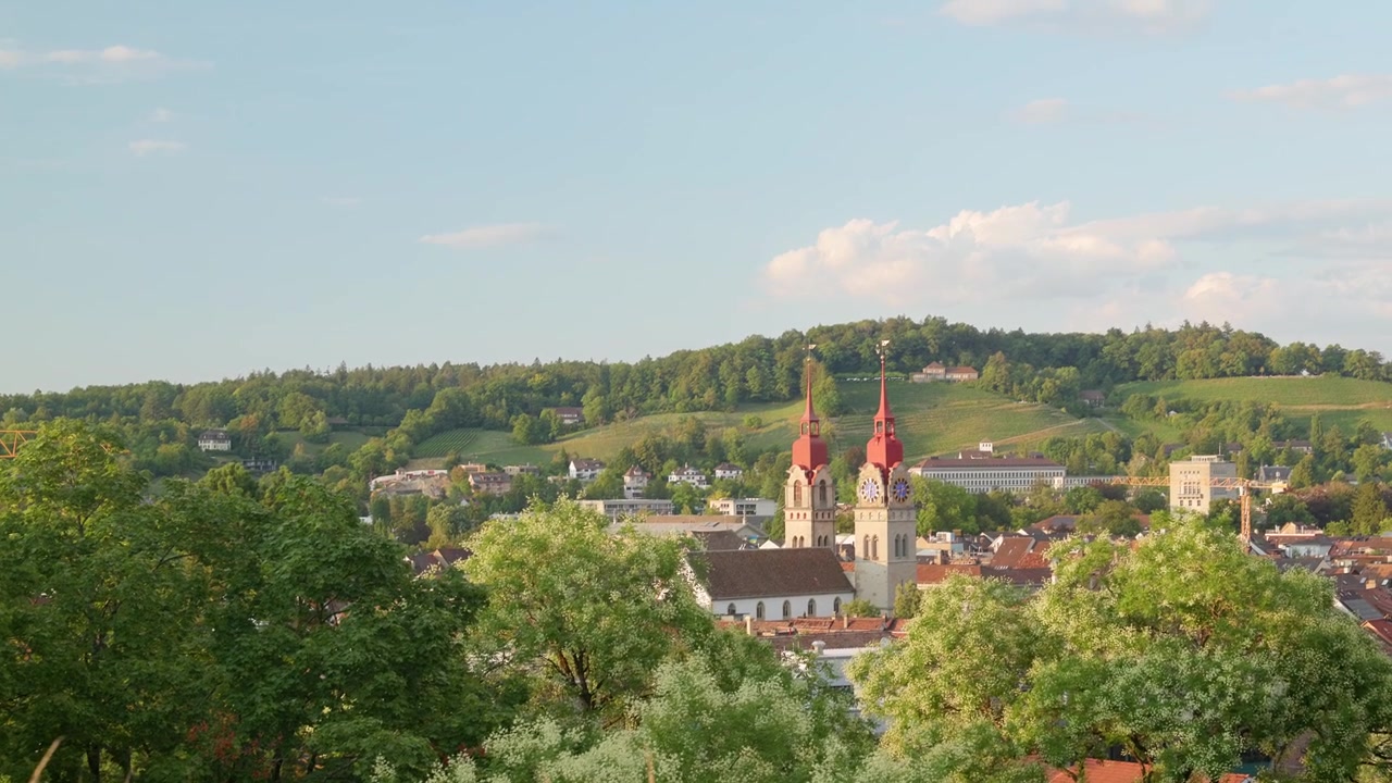Una vista panoramica di una cittadina immersa tra colline verdi, dominata dagli imponenti campanili rossi di una chiesa. In primo piano si estendono alberi rigogliosi, alcuni in fiore