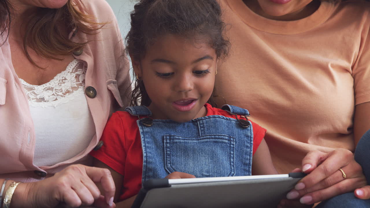 Latinamerikansk kvindefamilie på tværs af generationer sidder i sofaen derhjemme og bruger en digital tablet