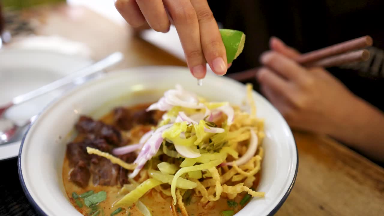 A hand squeezes fresh lime over a bowl of Northern Thai beef curry noodles at a wooden table, with natural lighting and shallow depth of field