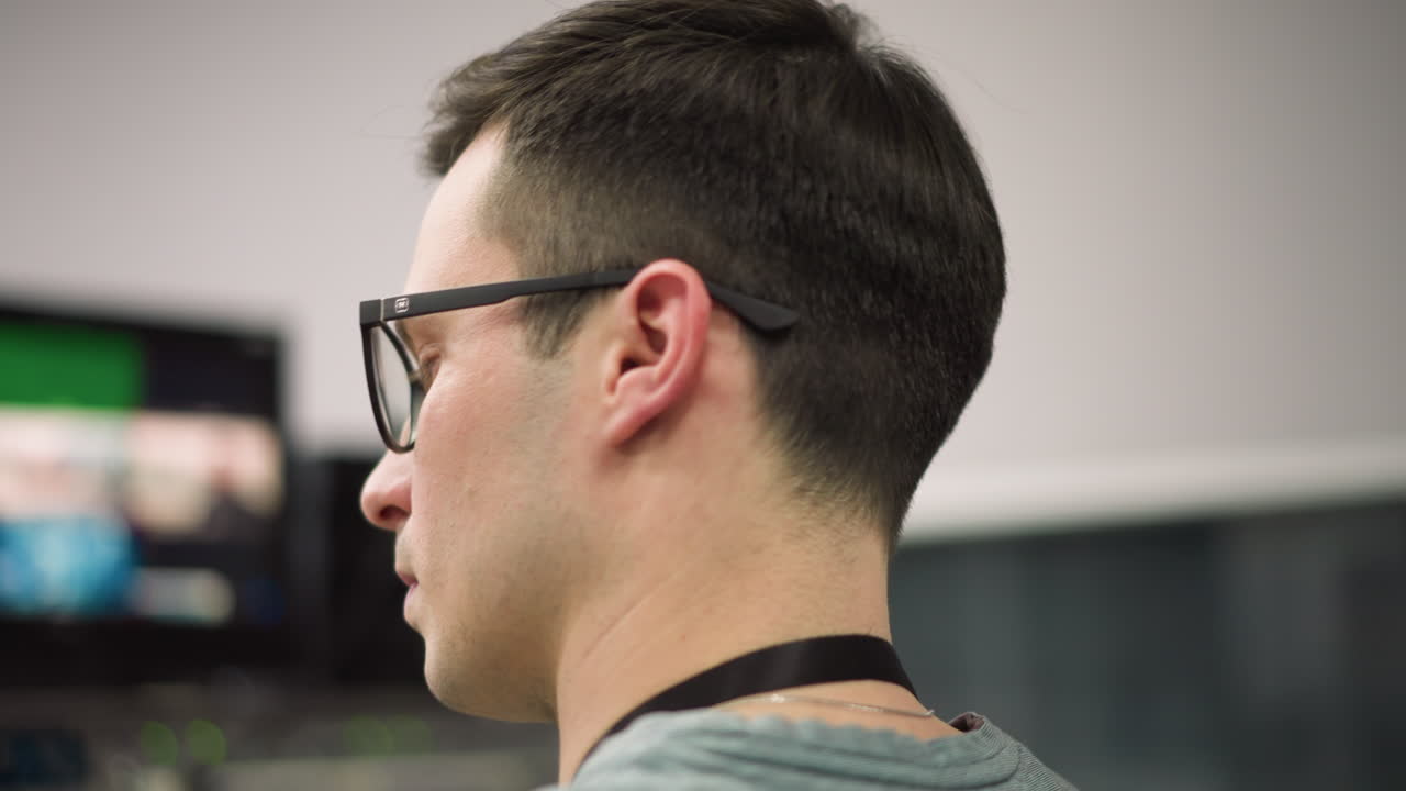 Close-up of young man with glasses, focused and deep in thought, working in office or studio environment. Profile shot showing concentration and professionalism in tech-related task