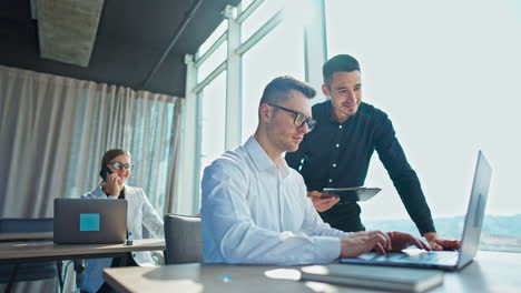 two young males look at the screen of laptop in the office. female colleague at desk behind chatting on the phone cheerfully.