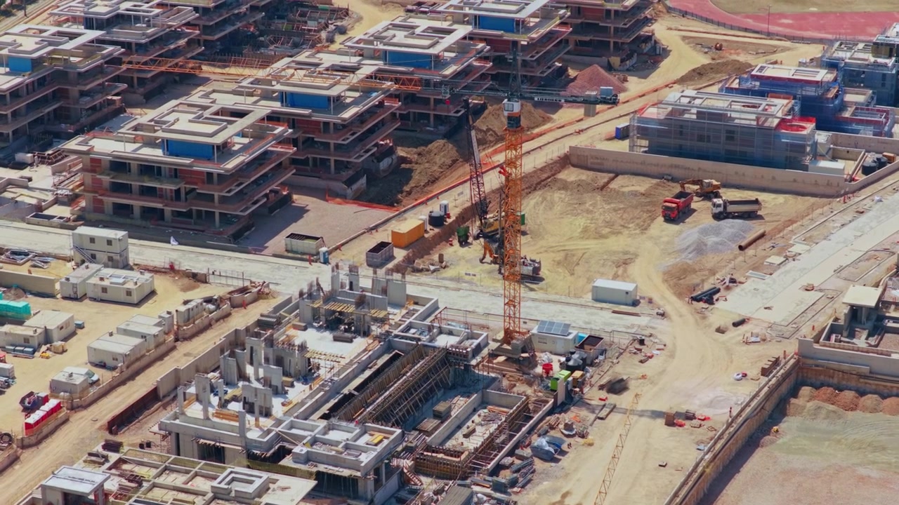 Amplia obra de construcción con múltiples edificios residenciales en diversas etapas de desarrollo, con grúas torre y maquinaria pesada operando en el suelo en un nuevo barrio urbano