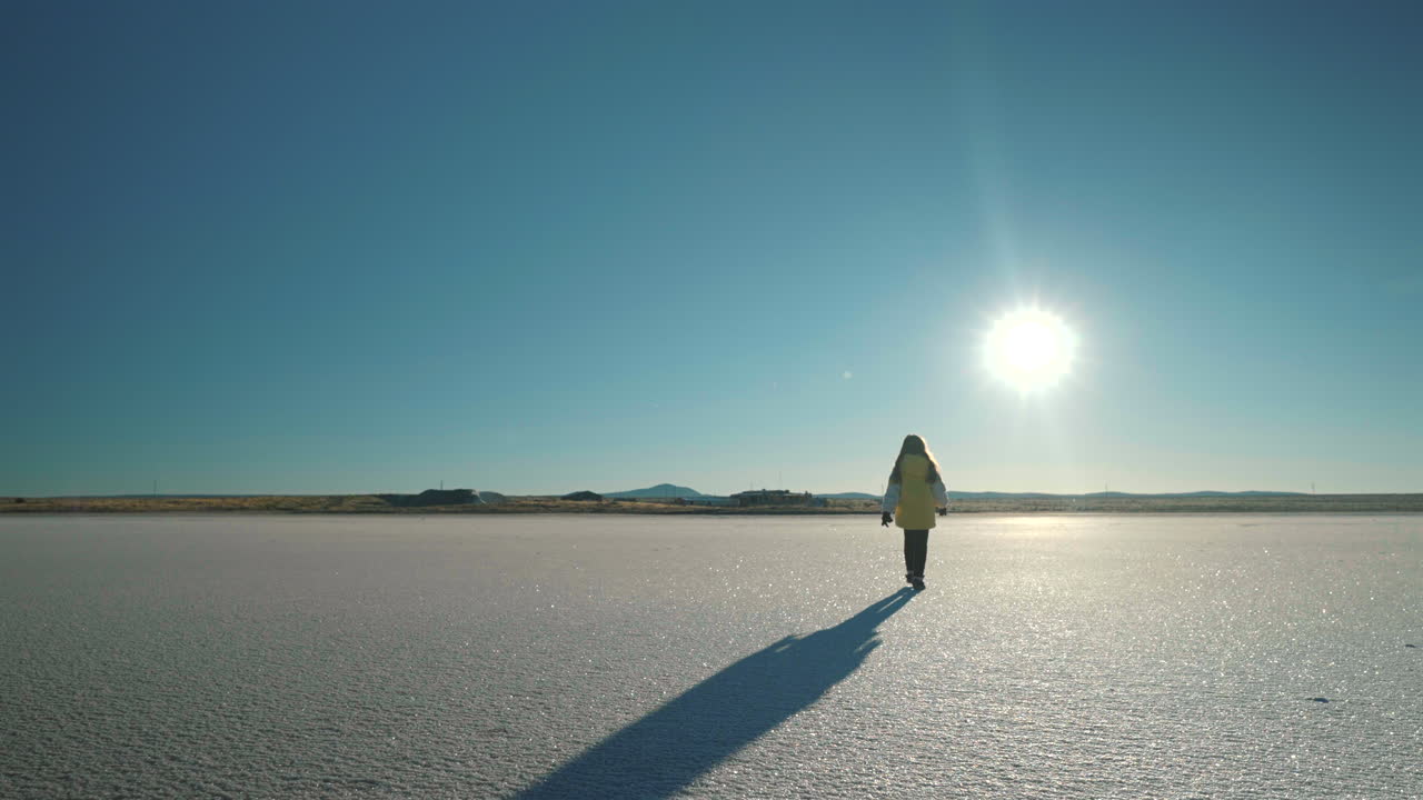 Slow motion static of girl in yellow jacket walking away across Salinas del Diamante expanse under intense sun, backwards silhouette diminishing into vast white salt pan near San Rafael, Mendoza