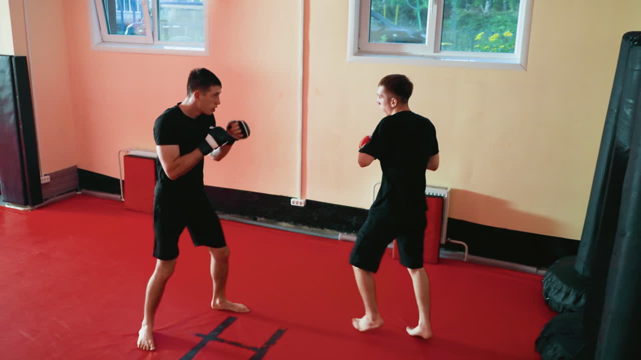 Wrestlers punching and kicking inside training gym, wearing black sportswear and gloves, standing barefoot on red mat, maintaining defensive stance with focused expressions