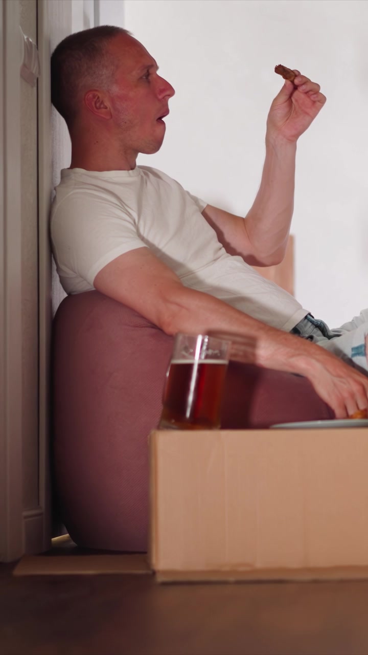 Primo piano di un uomo seduto su un pouf rosa che mangia uno spuntino con una bevanda in bicchiere in una scatola di cartone nelle vicinanze. L'uomo allunga la mano per prendere il cibo e lo mangia mentre è seduto contro il muro. Ambiente interno con illuminazione soffusa.
