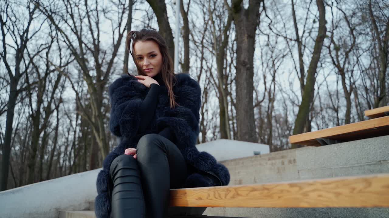 Beautiful girl in a coat sitting on a bench in a city park. Portrait of a luxury young woman in fur coat looking at camera and beckon with hands to herself. Slow motion.