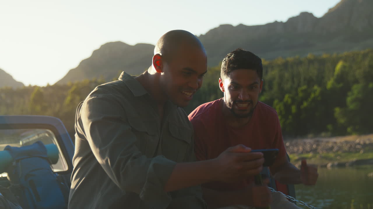 Zwei männliche Freunde mit Rucksäcken in einem Pickup-Truck am See, die Bier trinken und Sport auf dem Handy schauen