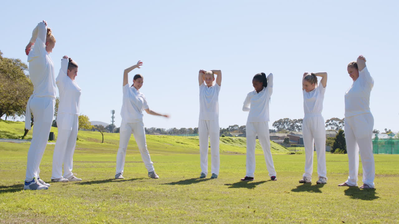 Women in white uniforms stretching together on cricket field, preparing for game
