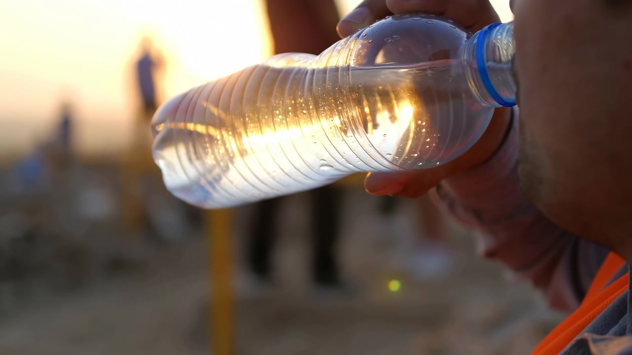 Una mano stabilizza una bottiglia parzialmente piena mentre una persona beve, con la linea dell'acqua inclinata che cattura la retroilluminazione dorata attraverso la plastica trasparente su uno sfondo esterno leggermente sfocato
