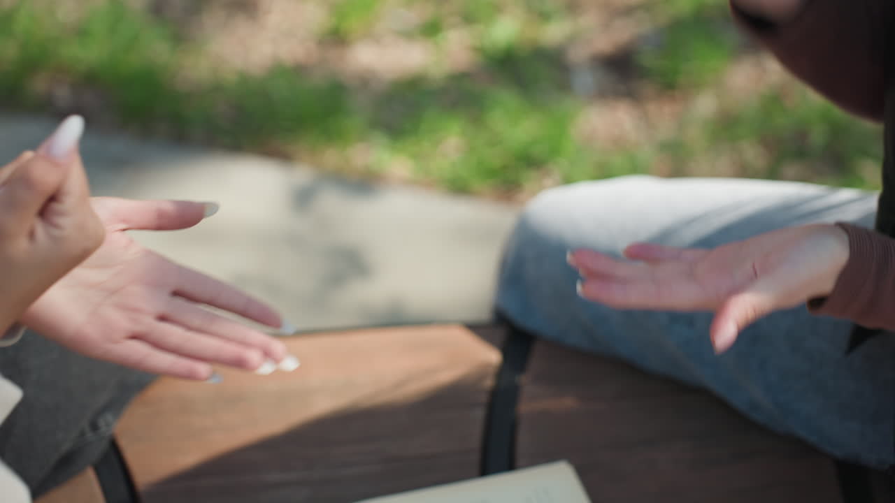 공원 벤치에서 손뼉 치기 게임을 하는 학생들, 백인 여성과 남성이 나무 테이블 위의 책과 휴대폰 위에서 손으로 숫자를 세는 제스처를 보여주는 모습, 햇빛이 비치는 캠퍼스 분위기, 얕은 피사계 심도, 솔직하고 친근한 분위기