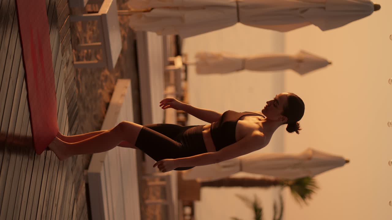 Vertical video: A brunette girl with tied hair in a black sports uniform lays out a red yoga mat and prepares for a yoga class on a sunny beach in the morning
