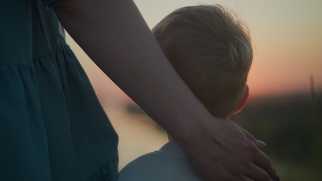 A tender close-up capturing a mother gently rubbing her young son's head as they both watch a serene sunset over a tranquil lake. The warm, golden light of dusk envelops them