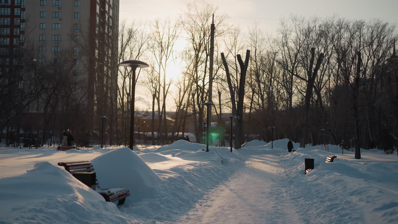 Serene winter scene with glowing sunlight casting warm hue over snow covered benches, trees, and street lamps along quiet park path lined with people walking and distant buildings in soft focus
