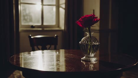 sunlight is streaming through the window and illuminating a red rose in a crystal vase sitting on a round wooden table in a dark room, creating a nostalgic and romantic atmosphere