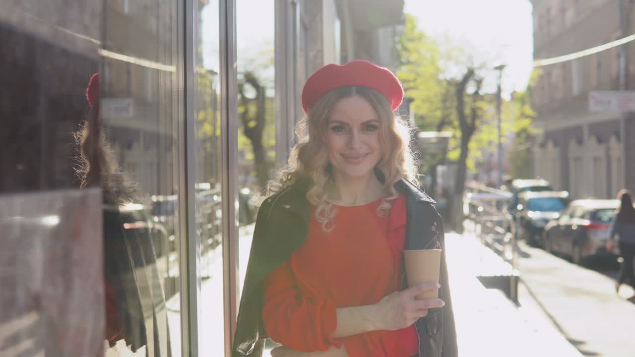 elegante joven rubia en un vestido rojo y trenza negra se encuentra en una calle acogedora en el sol de la mañana