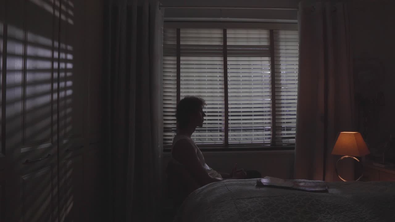 Man sits alone in moody room with blinds casting shadows, slow quiet motion, introspective