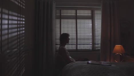 man sits alone in moody room with blinds casting shadows, slow quiet motion, introspective