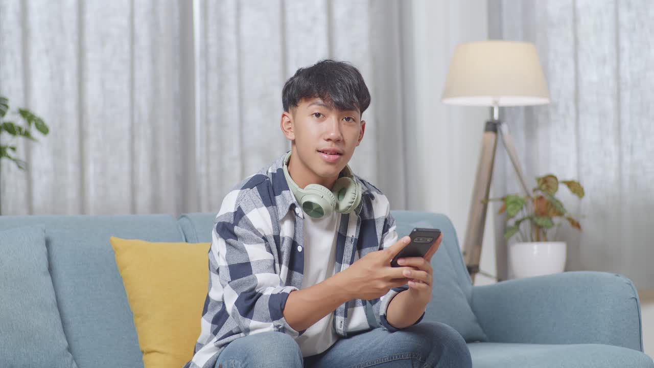 집 의 거실 의 소파 에서 스마트폰 을 재생 하는 동안 헤드폰 을 들고 카메라 를 향해 미소 짓는 아시아 청소년 소년
