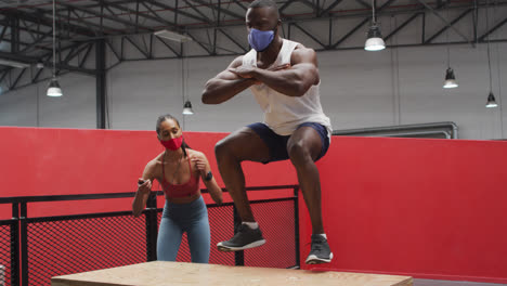 Hombre-Afroamericano-Saltando-Sobre-Una-Caja-Usando-Máscara-Facial-En-El-Gimnasio,-Mujer-De-Raza-Mixta-Sincronizada