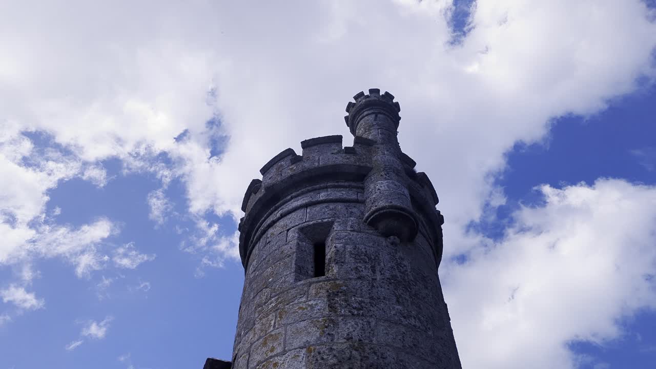 A low-angle shot looking up at a stone tower of Monterreal Castle in Baiona, Galicia, Spain. Fast-moving clouds pass over the medieval fortress battlements against a vivid blue sky.