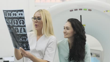 woman doctor radiologist explains the results of ct scanning for young female patient, showing the snapshot with images, observing and analyzing ct scan in modern clinic beside modern ct scanner