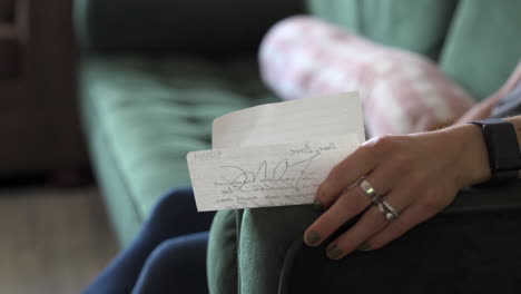 a close-up shot of a woman’s hand gently holding a love letter while she sits on a green couch. the camera slowly moves around her hand, highlighting the intimate moment.