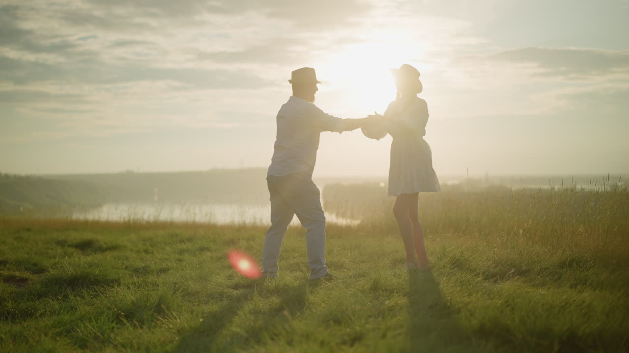 사랑하는 부부는 평화로운 호수 에 빛이 빛나는 잔디 언덕에서 춤을 춘다. 색 셔츠, 모자, 청바지를 입은 남자는 색 드레스와 검은색 모자를 입은 아내와 함께