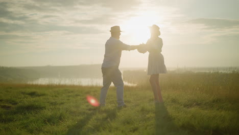 una pareja amorosa baila en una colina cubierta de hierba iluminada por el sol al lado de un lago pacífico. el hombre, vestido con una camisa blanca, sombrero y vaqueros con su esposa, que lleva un vestido blanco y un sombrero negro