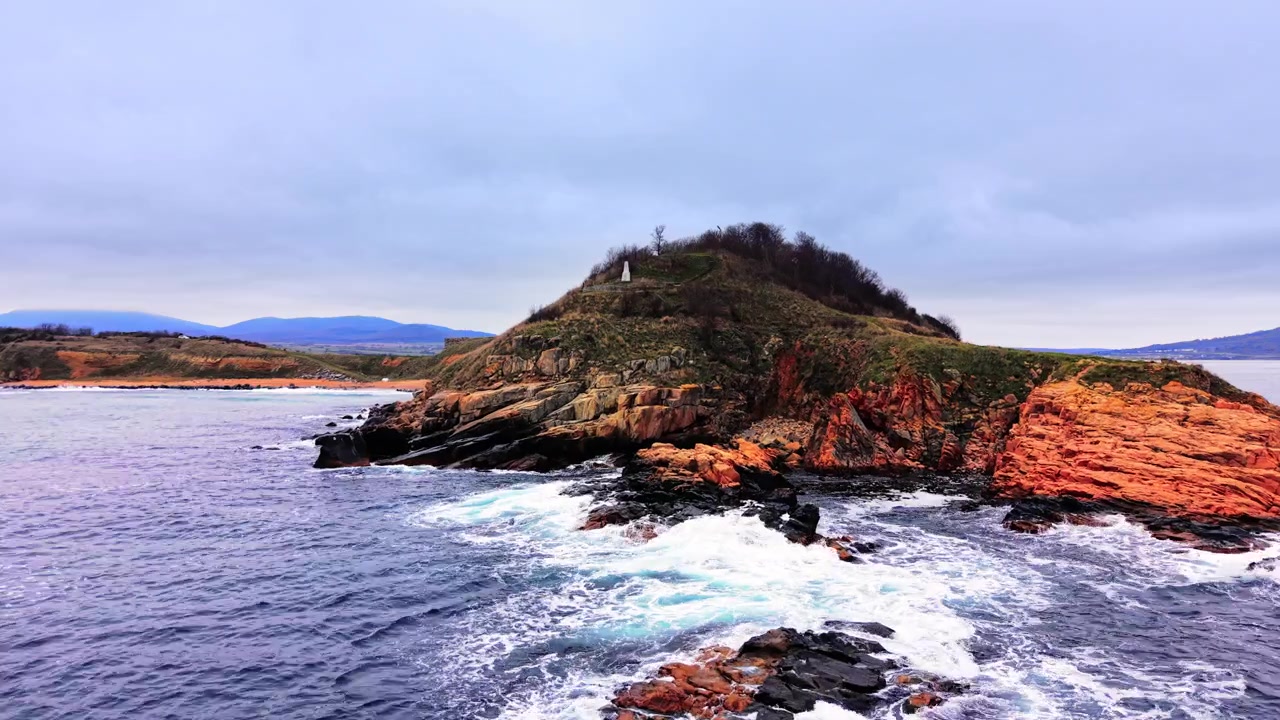 Rannikkonäkymä kallioisesta saaresta ja muistomerkistä, Sozopol, Bulgaria. Merenpinnan tasolta otettu kuvakulma näyttää vihreän kukkulan huipun, oranssin kallion ja tyynen veden pilvisen taivaan alla