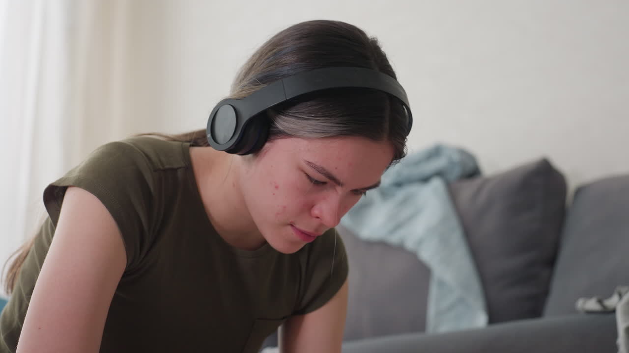 Side view of lady wearing headphones listening to music moving body gradually in relaxed rhythm while seated indoors near sofa with casual clothes around