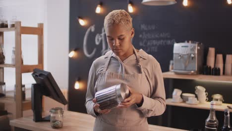 waitress with short hair looking at the camera while holding a glass jar full of coffee beans in a coffee shop