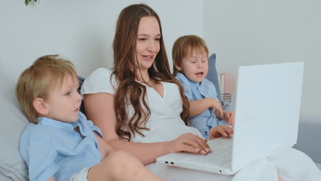 Beautiful-young-mom-and-two-little-kids-boys-are-looking-at-the-laptop-screen-family-photos.-And-they-do-online-shopping.-Call-grandmother-through-video-chat.-Children-show-their-fingers-on-the-laptop-screen-favorite-toys