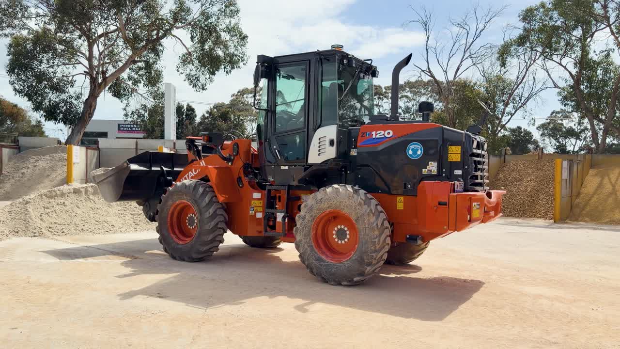 Orange front loader raises and lowers bucket on sandy construction site under bright daylight