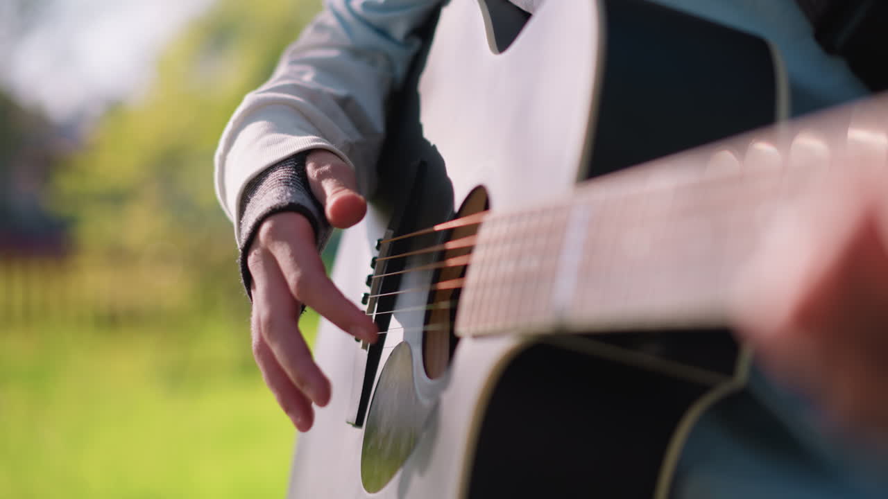 Caucasian guitarist plucking acoustic strings outdoors, bright afternoon sun, park greenery backdrop, closeup near soundhole and right hand, steady rhythmic strumming with pick, casual sweater